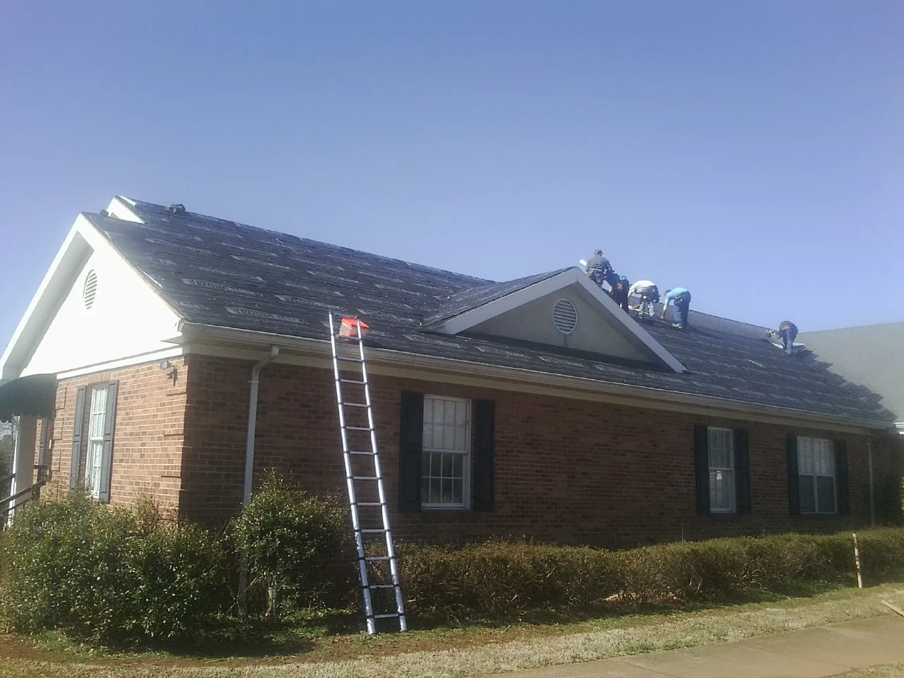 Crew tearing shingles off a brick ranch roof with a ladder set — Metal Roof Repair work by our crew in Le Roy