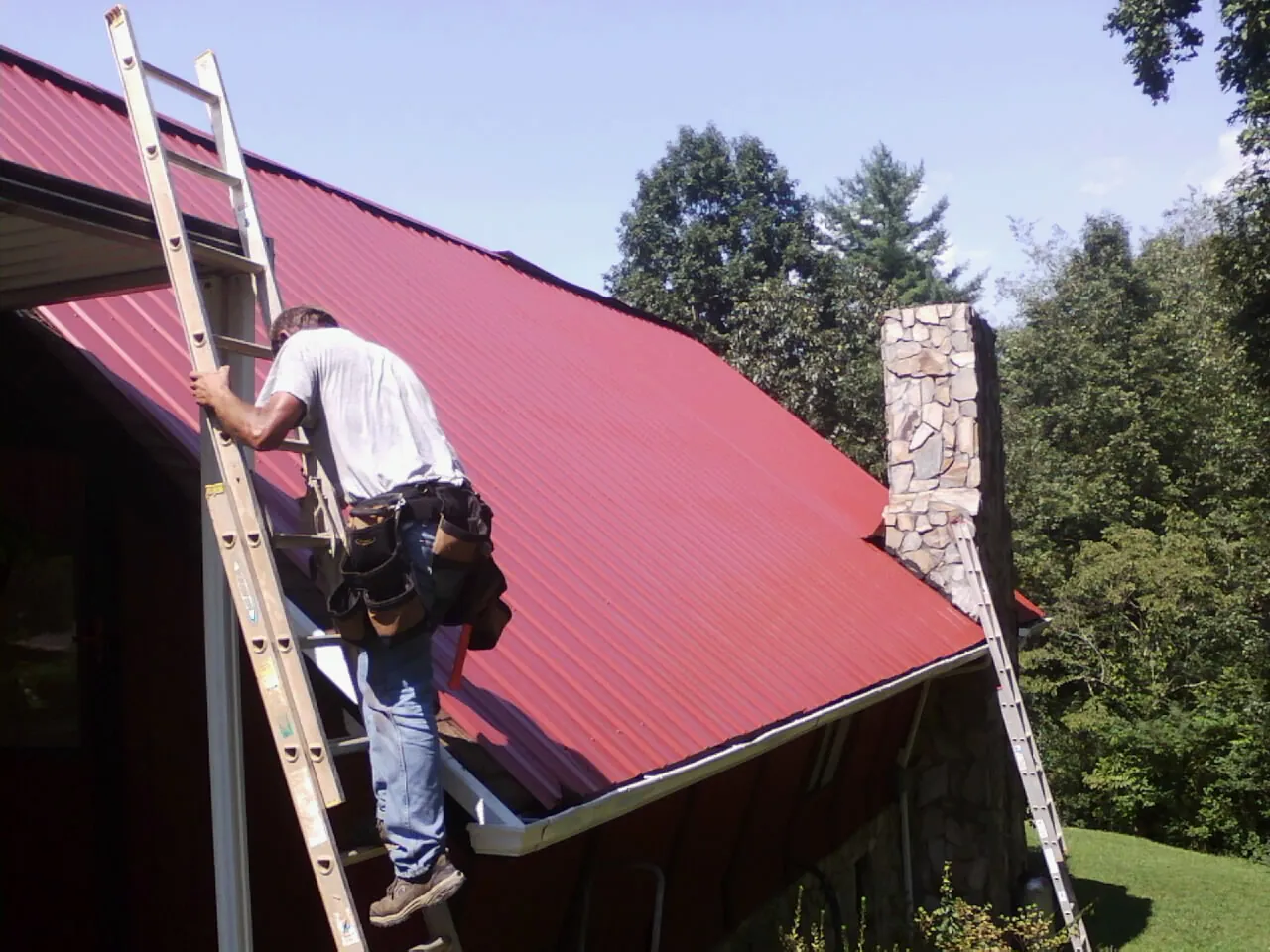 Roofer on a ladder beside a red metal roof and stone chimney — Soffit & Fascia Repair by Roofing Brothers in Le Roy