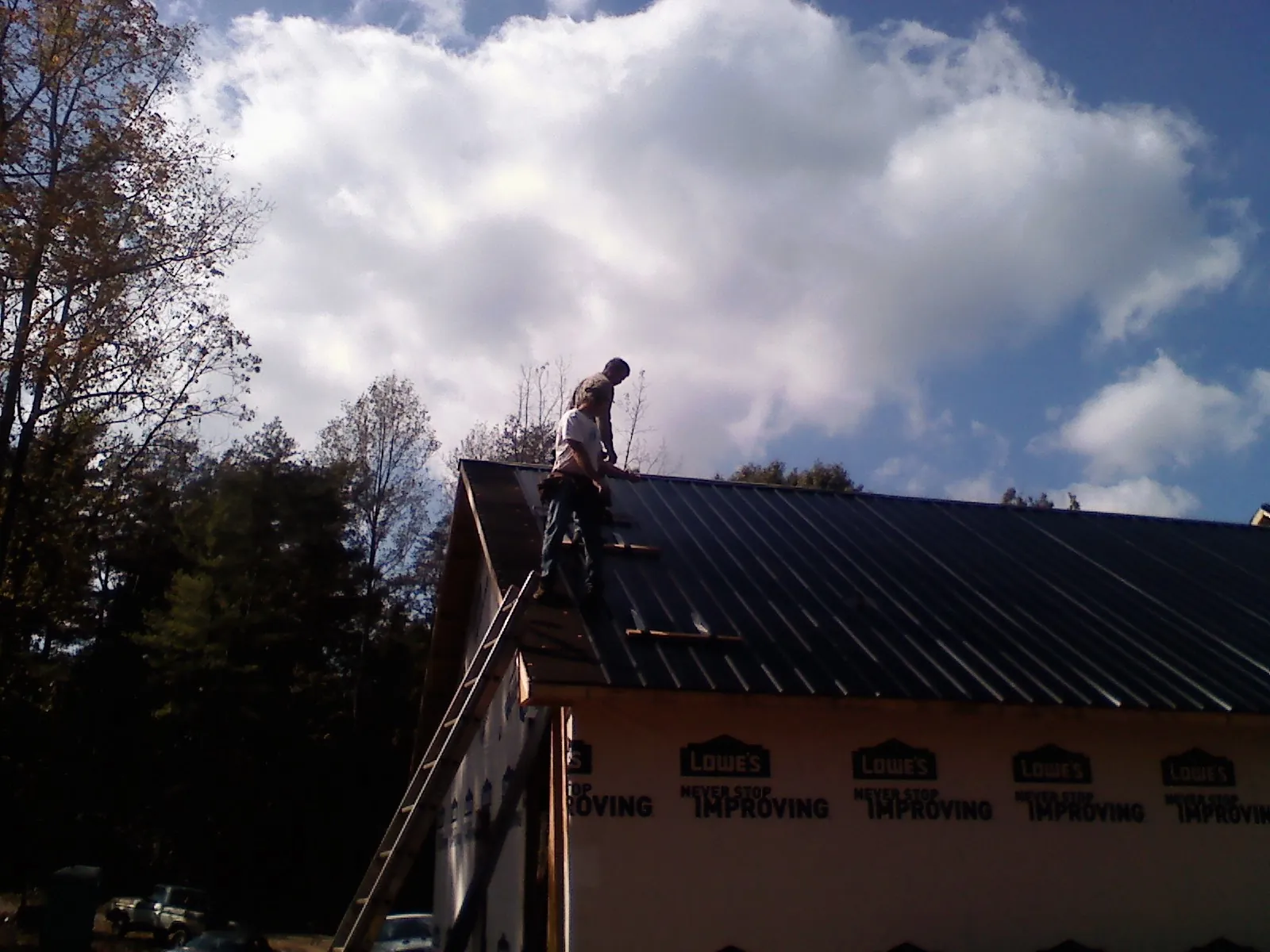 Roofer fastening dark metal panels high against a cloudy sky, from a Roof Inspection job in Le Roy